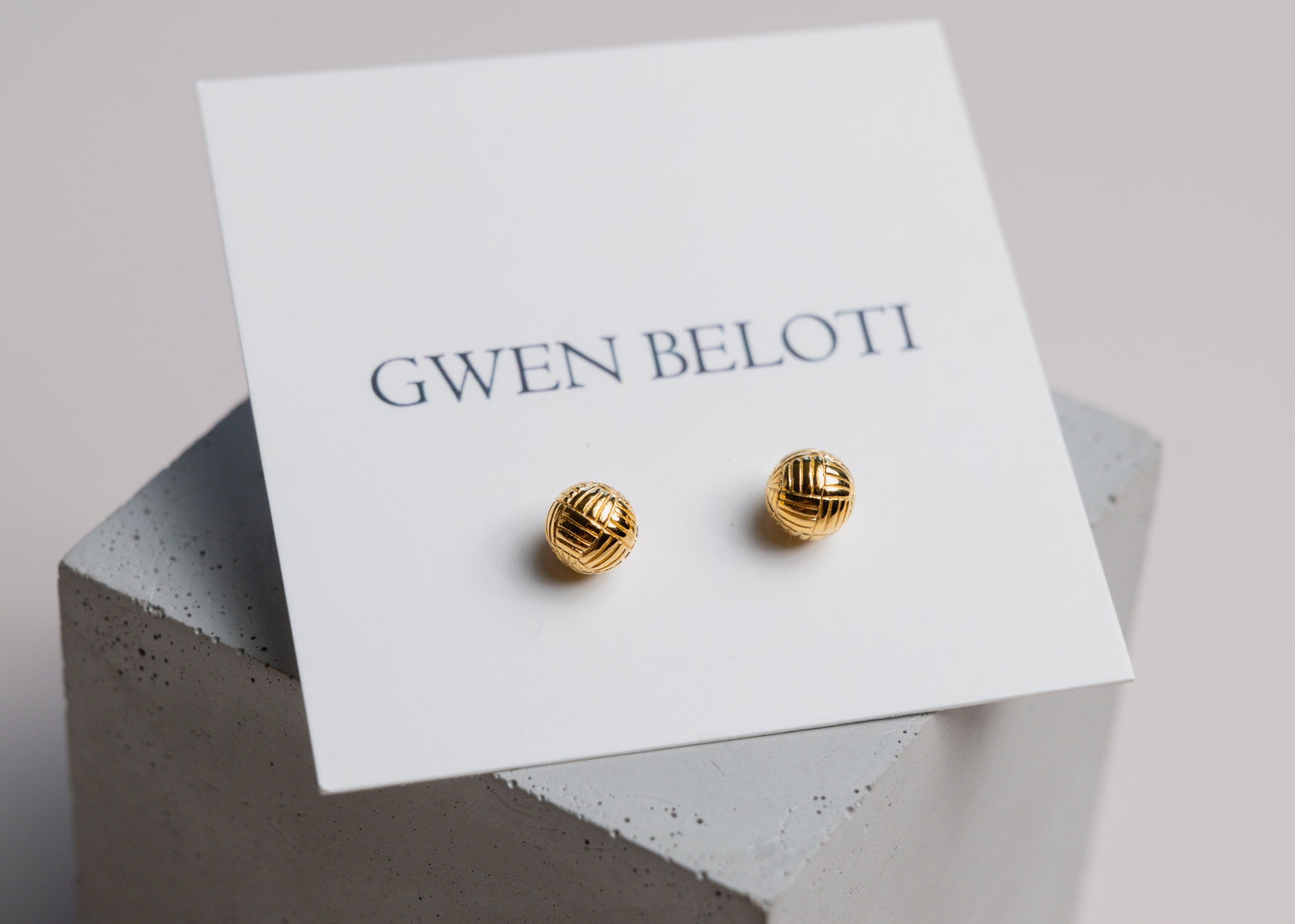 Gold woven ball earrings by Gwen Beloti displayed on a concrete block.