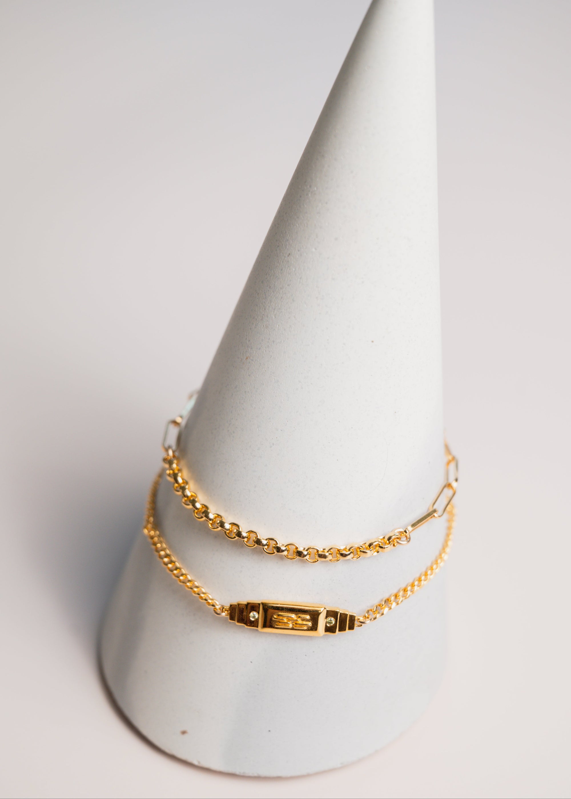 Two gold bracelets on a white cone-shaped stand against a light gray background