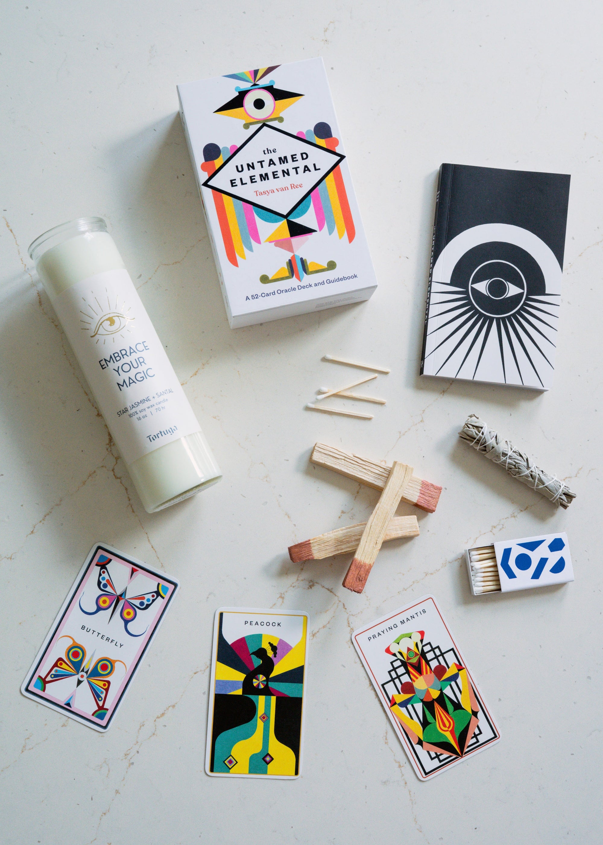 Collection of items including a prayer candle, oracle cards, palo santo, sage and matches on a light surface