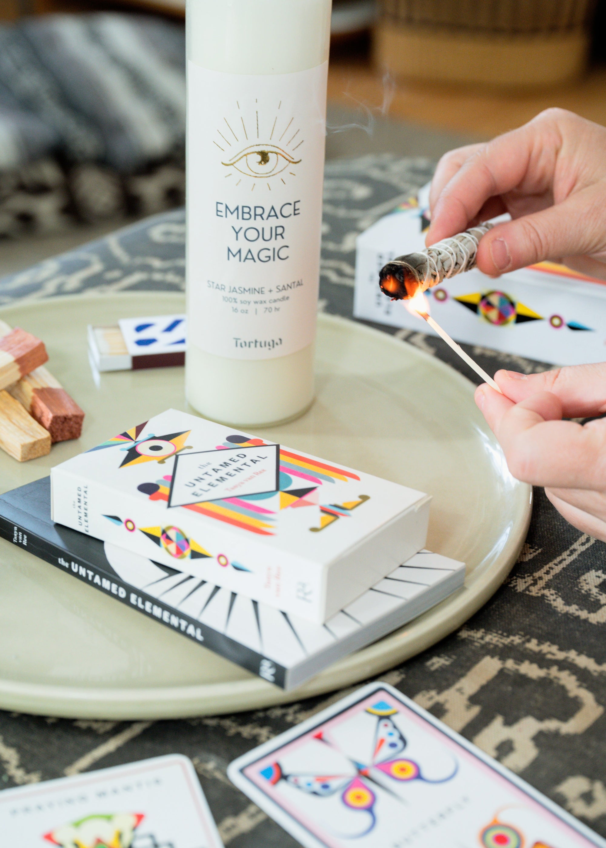 Person using lighting sage and a candle labeled 'Embrace Your Magic' with oracle cards and book on a table.