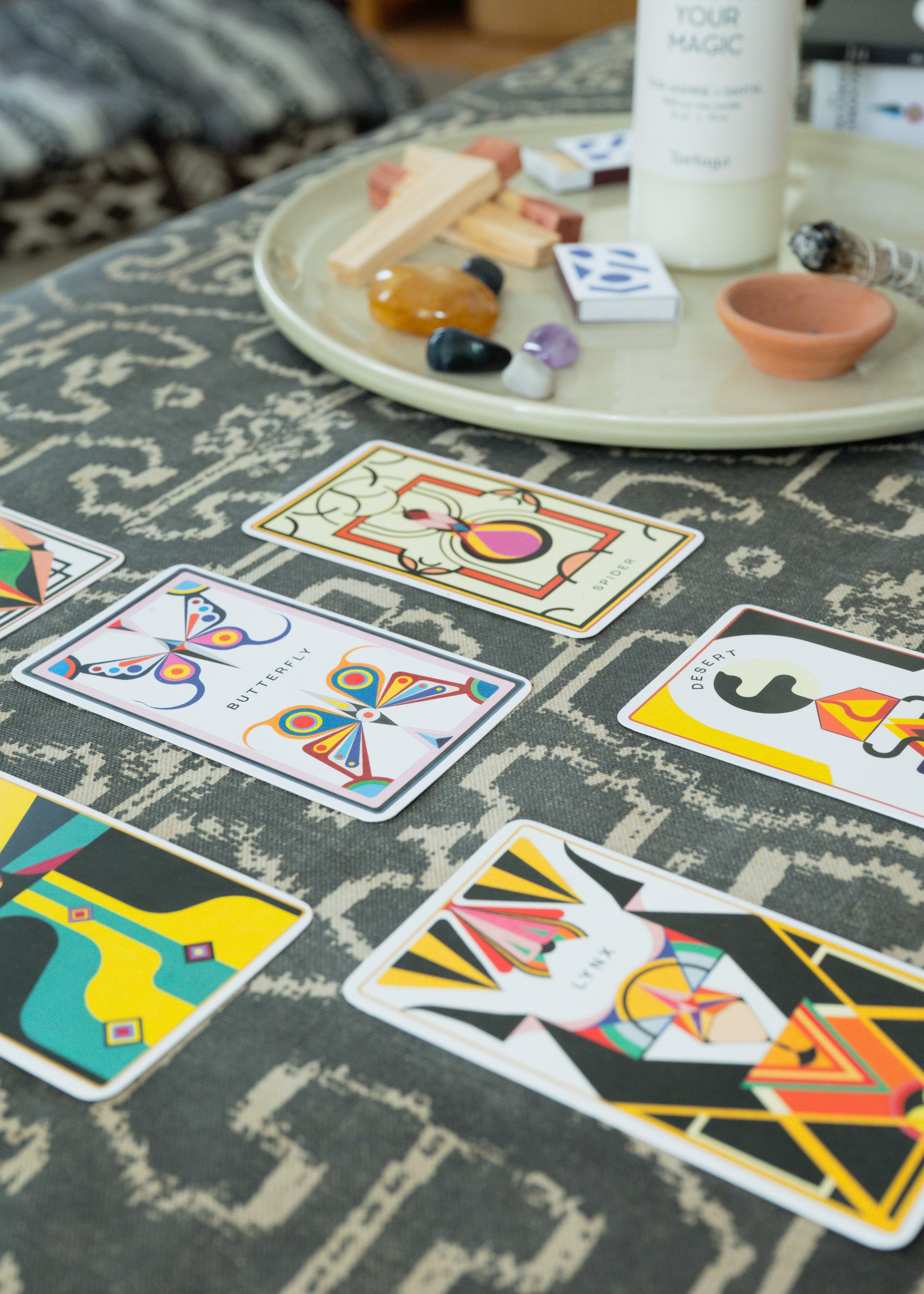 Colorful geometric oracle cards spread on a textured surface with a tray of crystals, sage, and palo santo.