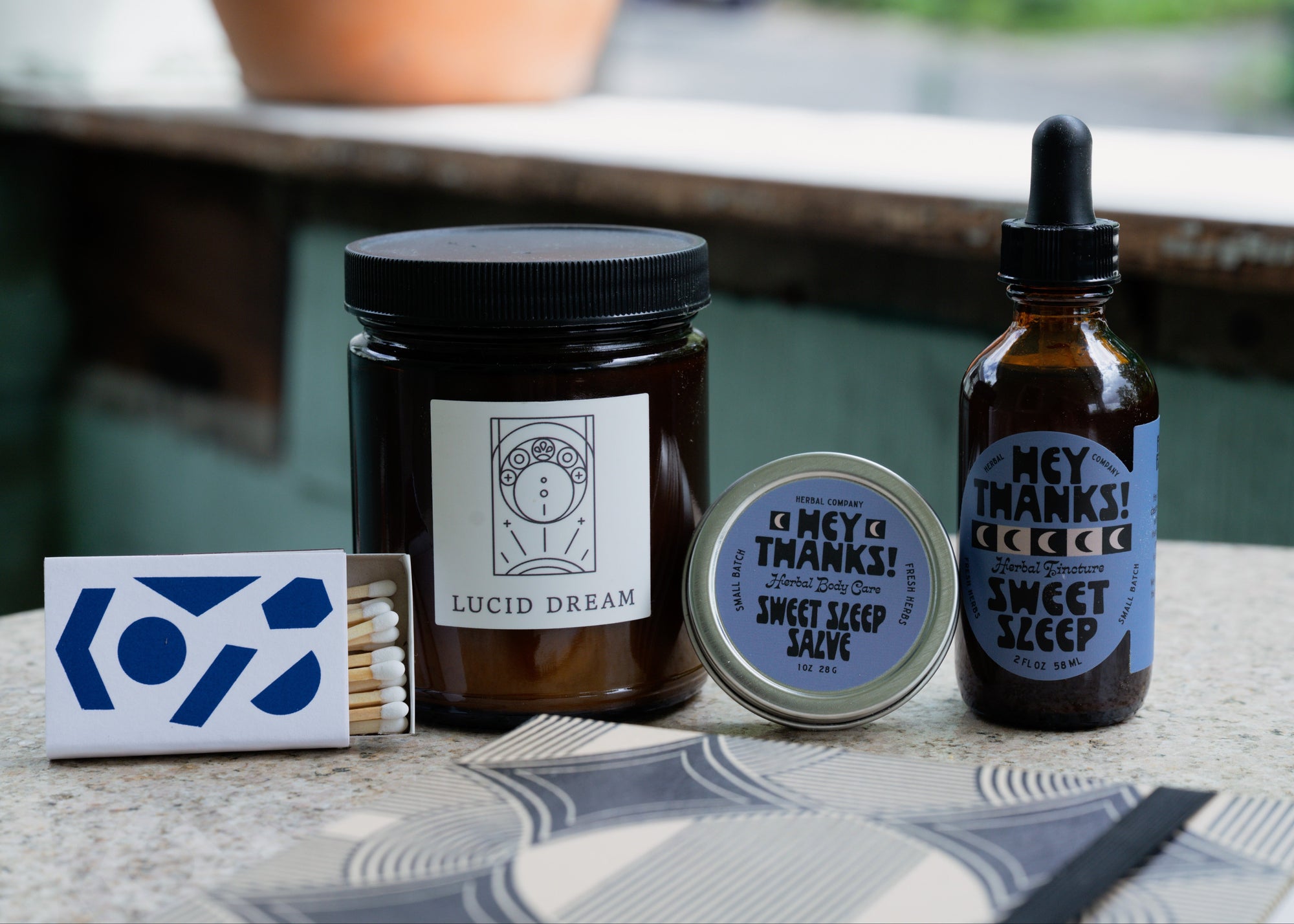 Collection of wellness products including a notebook, candle, tincture, salve, and matches on a table outdoors.