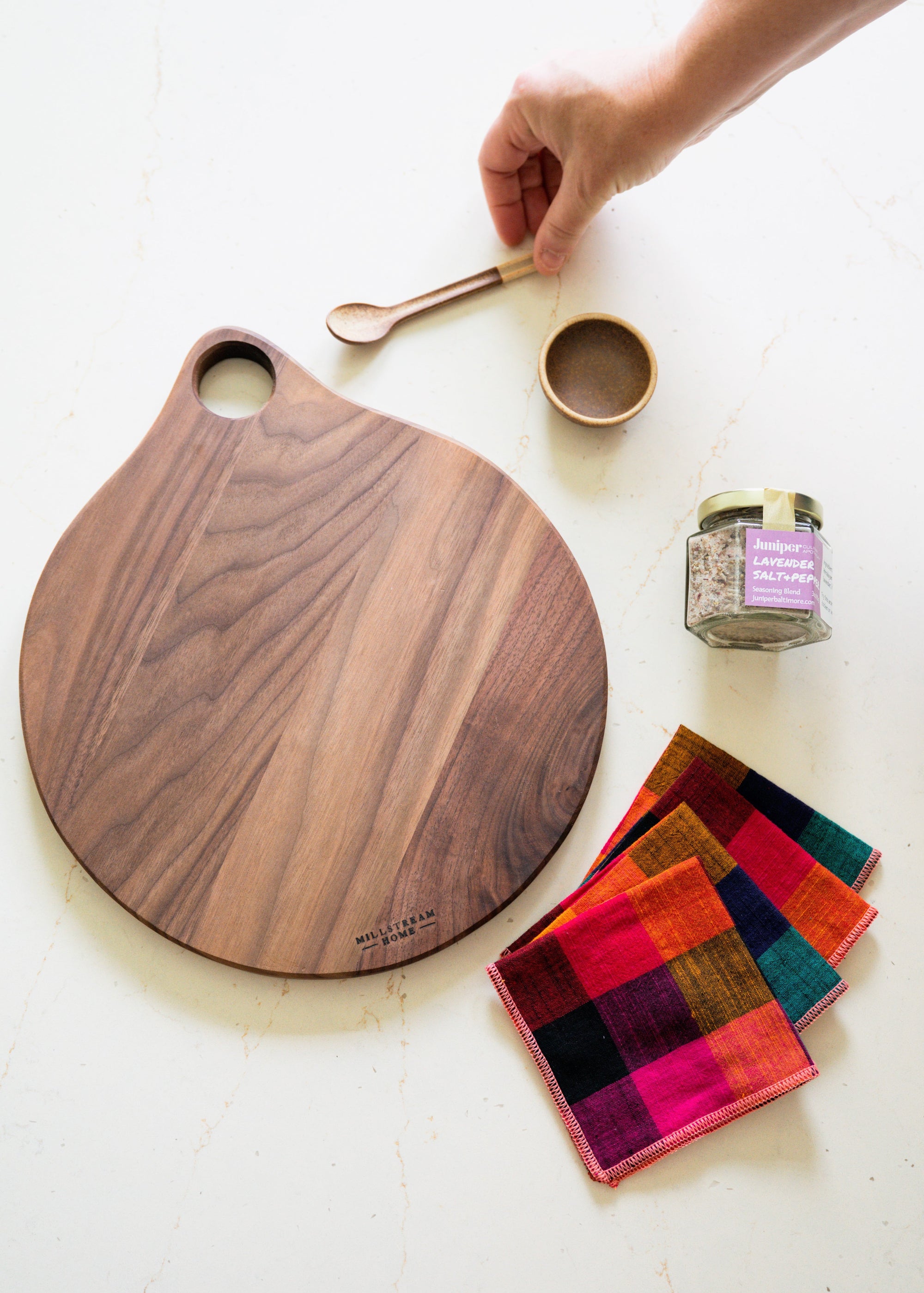 Wooden cutting board with colorful linen napkins, jar of salt, and a small ceramic bowl with spoon on a white surface