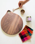 Wooden cutting board with colorful linen napkins, jar of salt, and a small ceramic bowl with spoon on a white surface