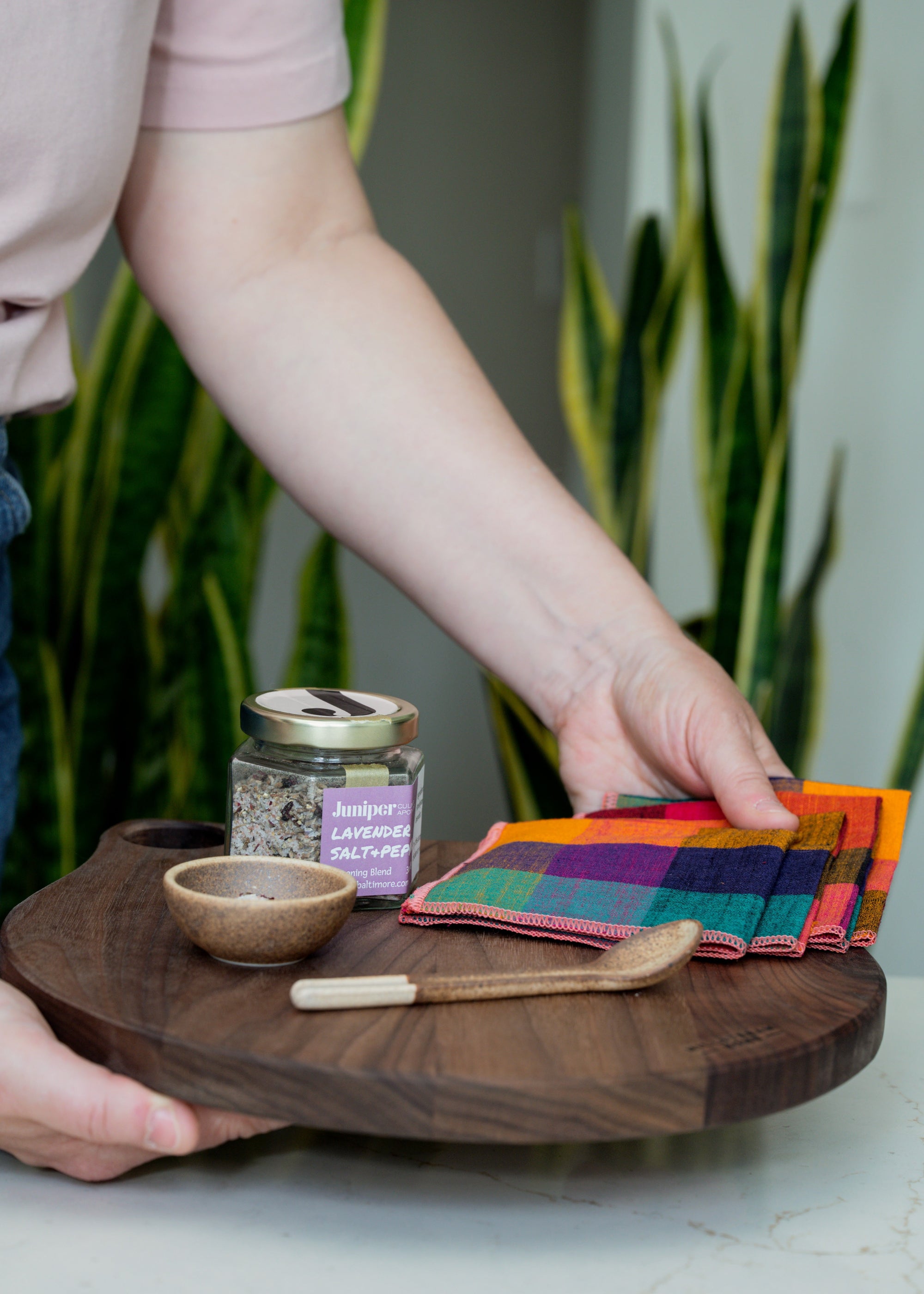Person holding a wooden serving board with colorful checkered napkins, a jar of salt and small ceramic bowl and spoon.