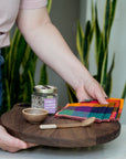 Person holding a wooden serving board with colorful checkered napkins, a jar of salt and small ceramic bowl and spoon.