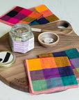 Colorful checkered napkins on a wooden serving board with a jar of salt and small bowl.