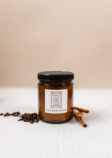 close up of a candle labeled "golden hour" with fresh cloves and cinnamon sticks besides the candle on a white background