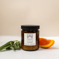 photo of a candle labeled "lucid dream" with orange halves and sage besides the candle on a white and light pink background