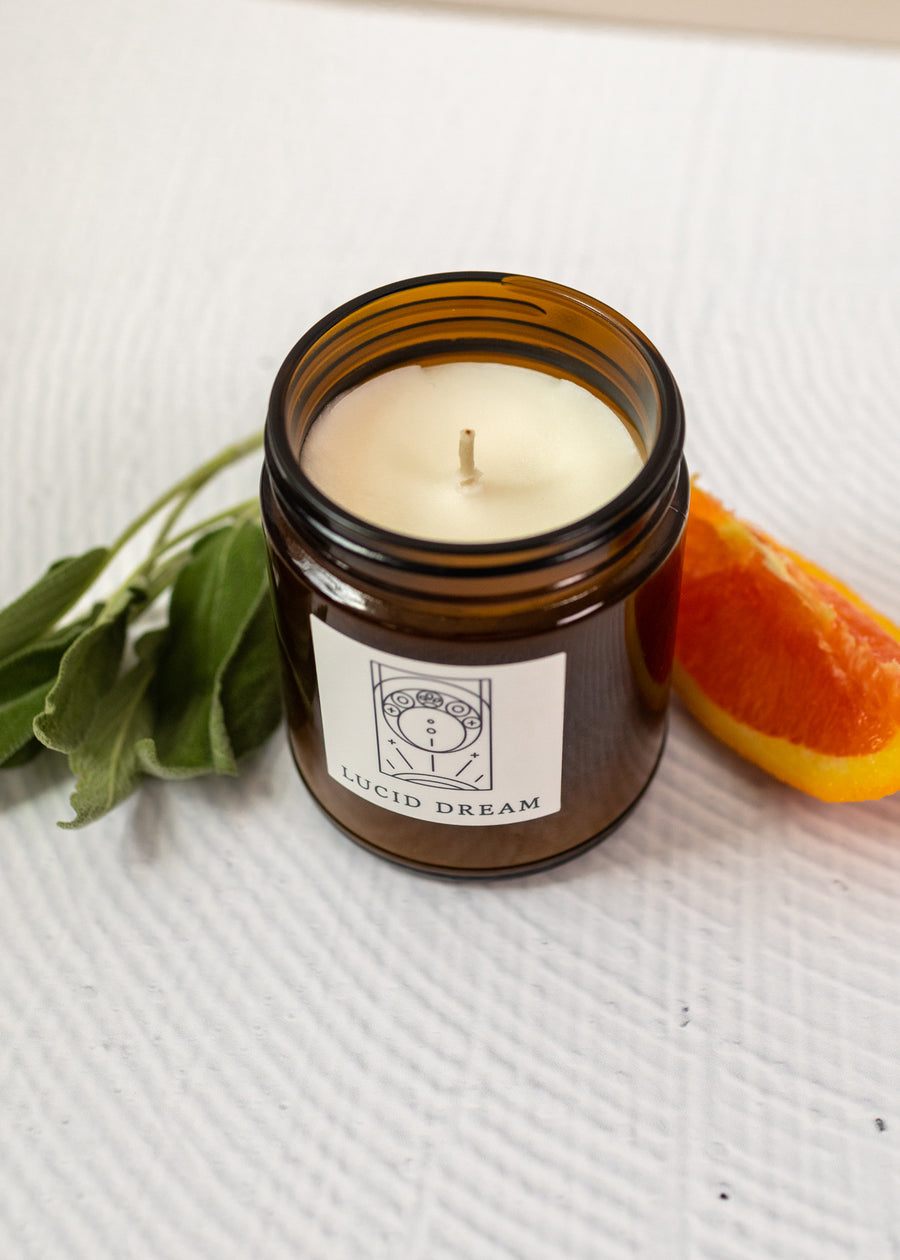close up of a candle labeled "lucid dream" with orange slices and sage besides the candle on a white background