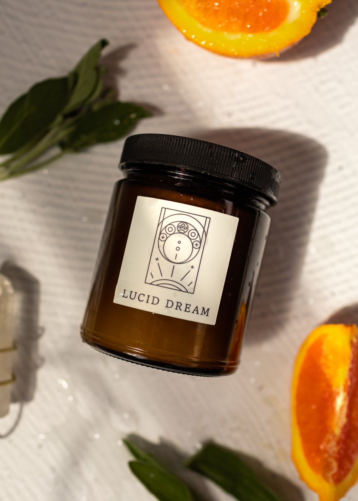 flat lay of a candle labeled &quot;lucid dream&quot; with orange halves and sage besides the candle on a white background