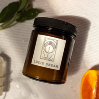 flat lay of a candle labeled "lucid dream" with orange halves and sage besides the candle on a white background