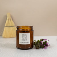 A candle labeled "sunset grasses" with a sprig of a flower next to it and a broom in the background.