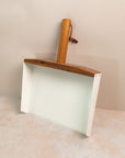 photo of a white large dust pan with a walnut handle, leaning against a pink background