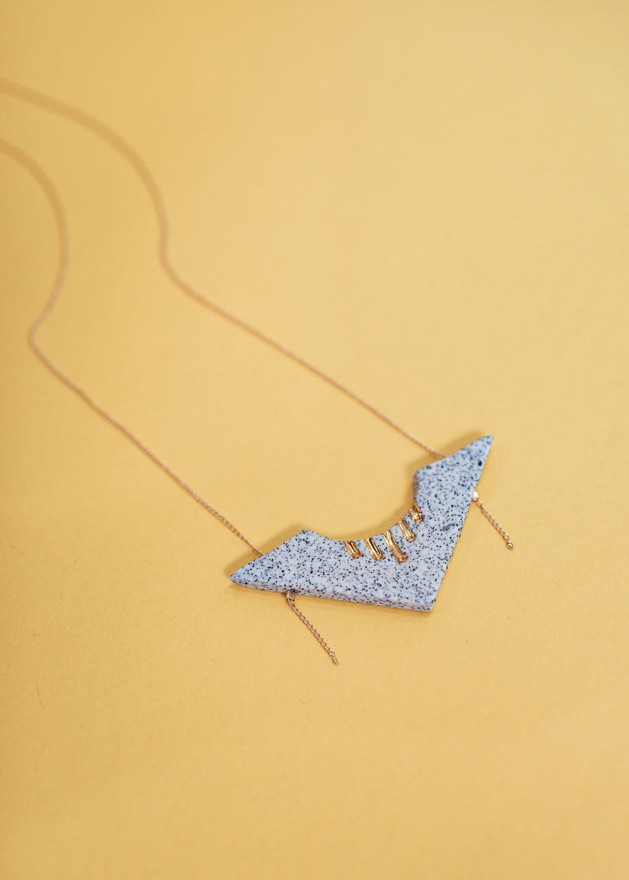 Photo of a necklace pendant made out of speckled granite. The granite is in the shape of an arrow, with 5 lines of gold at the top. Band attached to the necklace is dainty gold and extends through the arrow