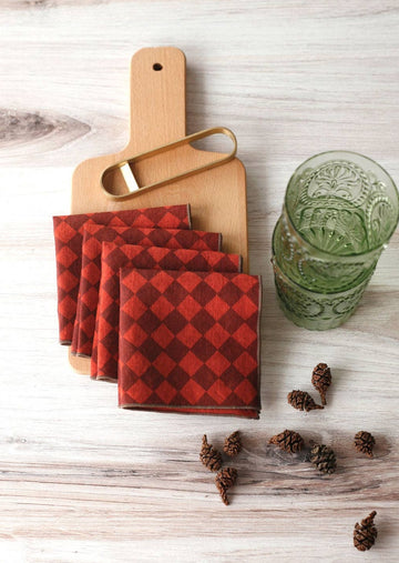 Four Diamond Cocktail Napkins in Ruby on a Serving Board with Pinecones surrounding and Green glasses to the right