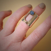 Long brass ring on a models upper half of finger with an arch that take up some of the ring. inside arch is a round Labradorite stone