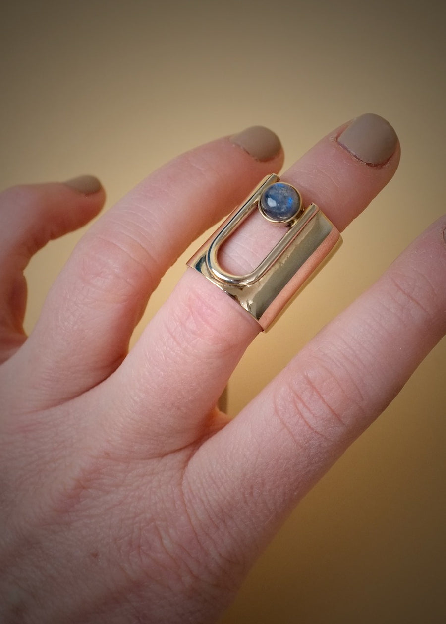 Long brass ring on a models upper half of finger with an arch that take up some of the ring. inside arch is a round Labradorite stone